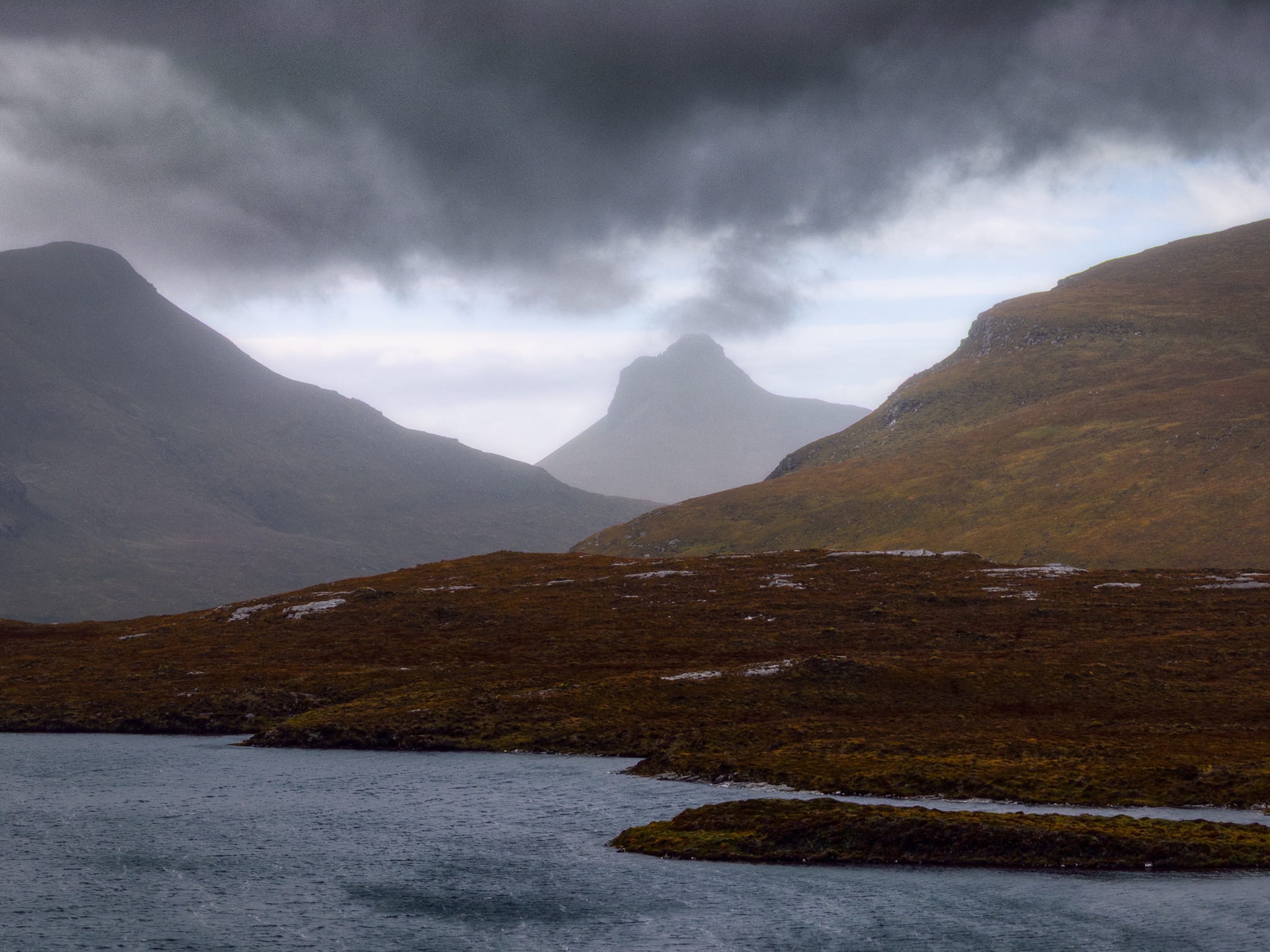 At the Knockan Crag Nature Reserve we take in the glorious sight across Lochan an Ais of Stac Pollaidh (612.4 m/2,009 ft), framed between the shoulders of Cùl Beag, (769 m/2,523 ft) and Cùl Mòr (849 m/2,785 ft). The driving storm clouds give Stac Pollaidh the impression that it&rsquo;s a volcano spewing ash into the sky.