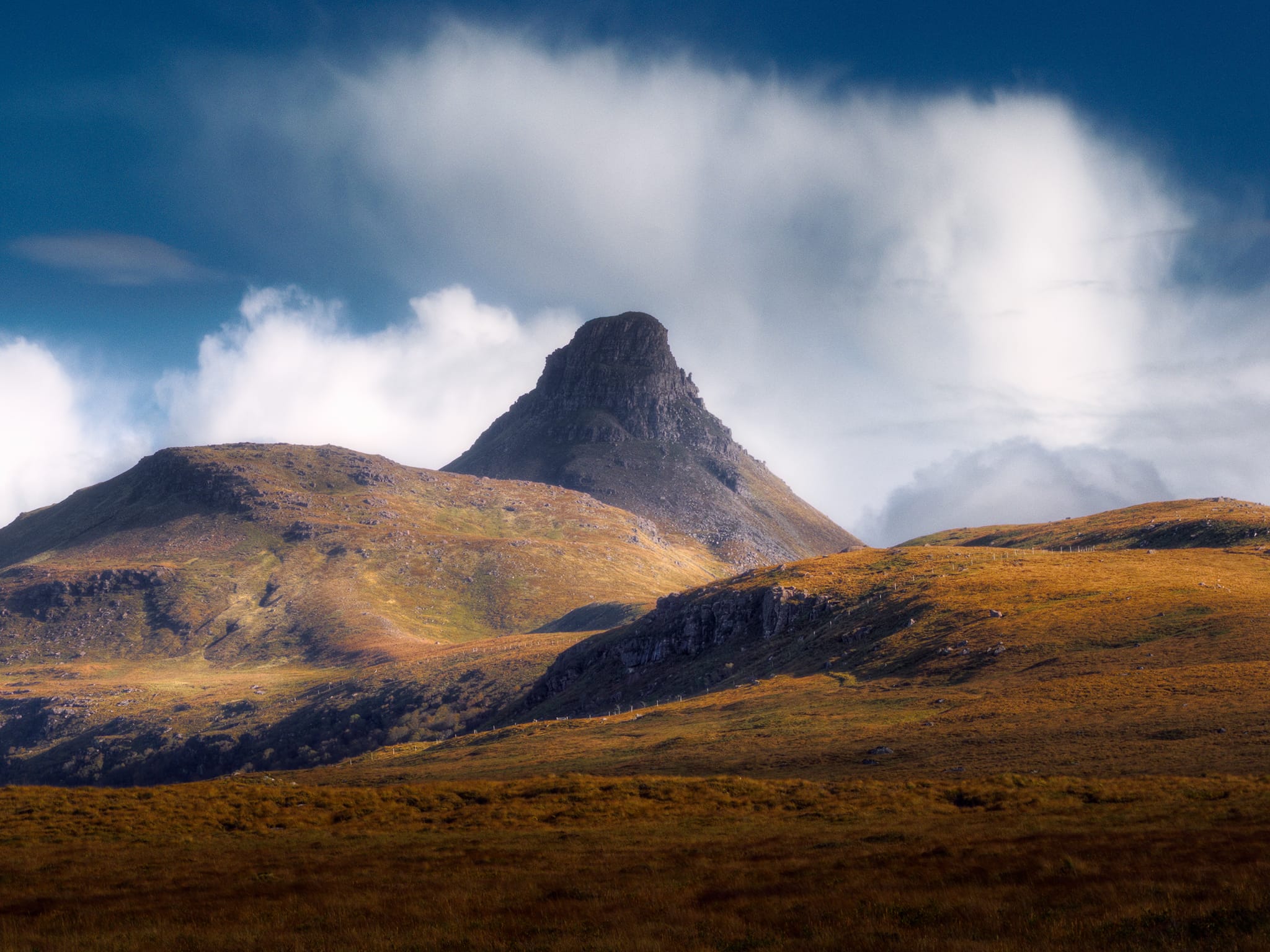 Further along the road the clouds were finally starting to move on. We drove past Loch Lurgainn then Loch Bad a&rsquo; Ghaill, taking the road at Badnagyle up and onto the moorland between Stac Pollaidh and Loch Garvie. It was here that I got my first side-profile view of Stac Pollaidh, looking like a slender pyramid topped with crags and pinnacles. The clouds broke apart and raced away, allowing light to pick out the beautiful contours of the landscape as I lined up this zoomed in composition of what was quickly becoming one of my favourite mountains.