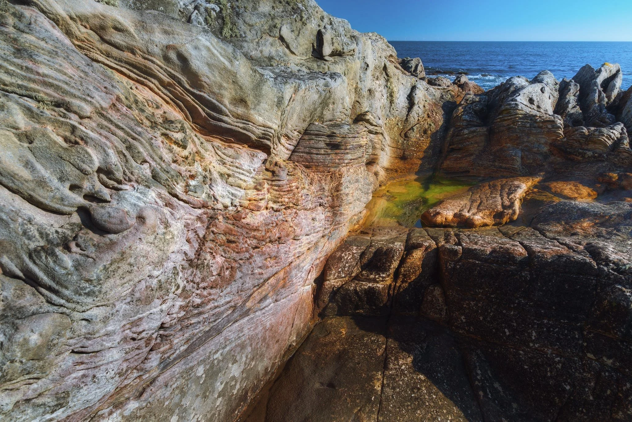  At Lochaber Rock, beautiful layers can be seen in the light-colored sandstone, with reddish-pink stripes. On the left side, you can see honeycomb-like patterns and wavy textures, which look different from the darker, sharp shapes on the right. These differences show how the rocks are made of different materials and react in unique ways to the ocean&rsquo;s wear. 