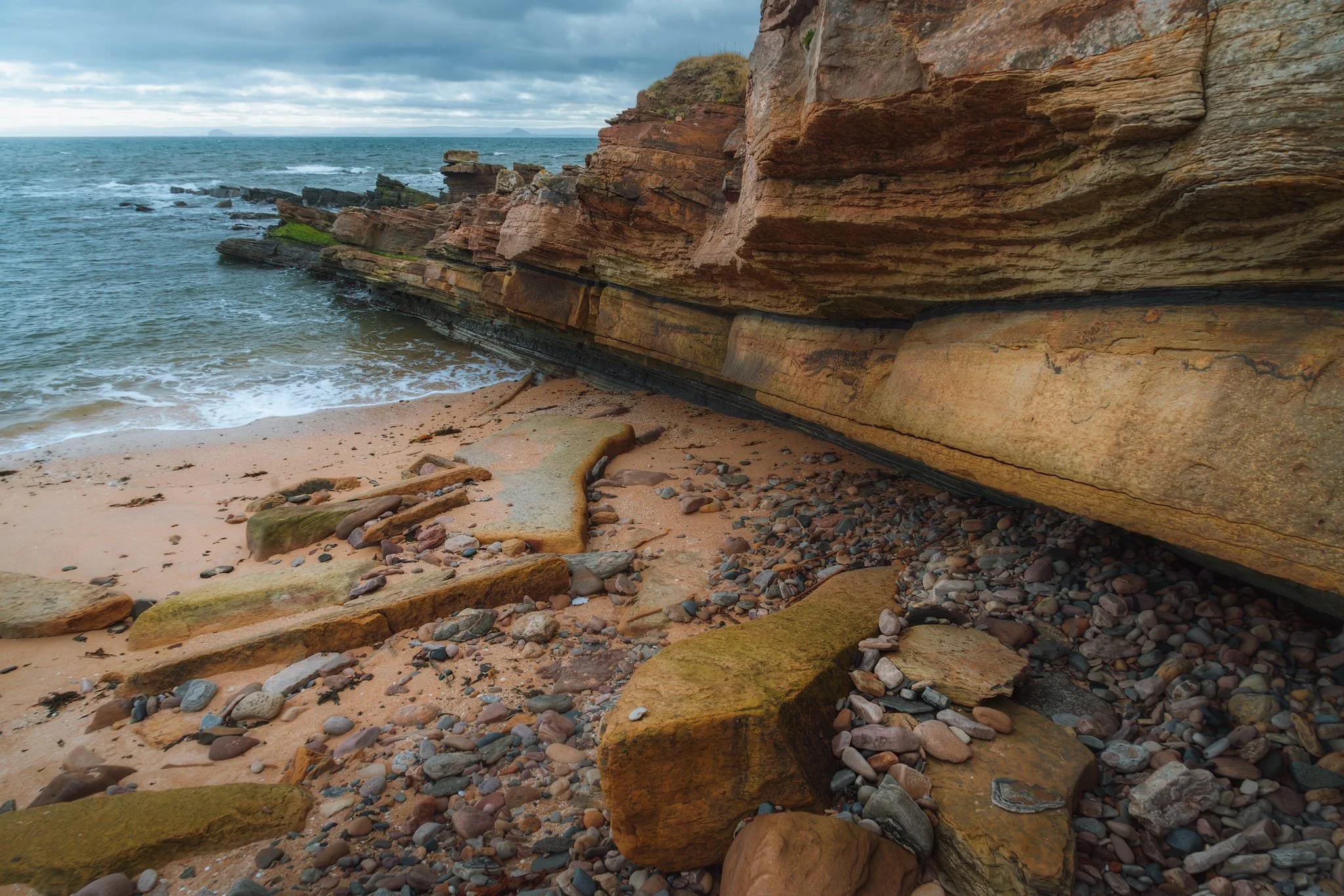  Below the playground and Anstruther Wester&rsquo;s golfing area, one can play around with the crazy rock formations at Billow Ness. The cliffs and boulders are primarily composed of sandstone; the layering indicates these rocks were deposited in horizontal strata, like an ancient river or sea. The reddish-brown colouring hints at the presence of iron oxides. In the distance, across the Firth of Forth, you can just about make out two distinct &ldquo;bumps&rdquo; on the horizon. The one on the left is Bass Rock, and on the right is Berwick Law. 