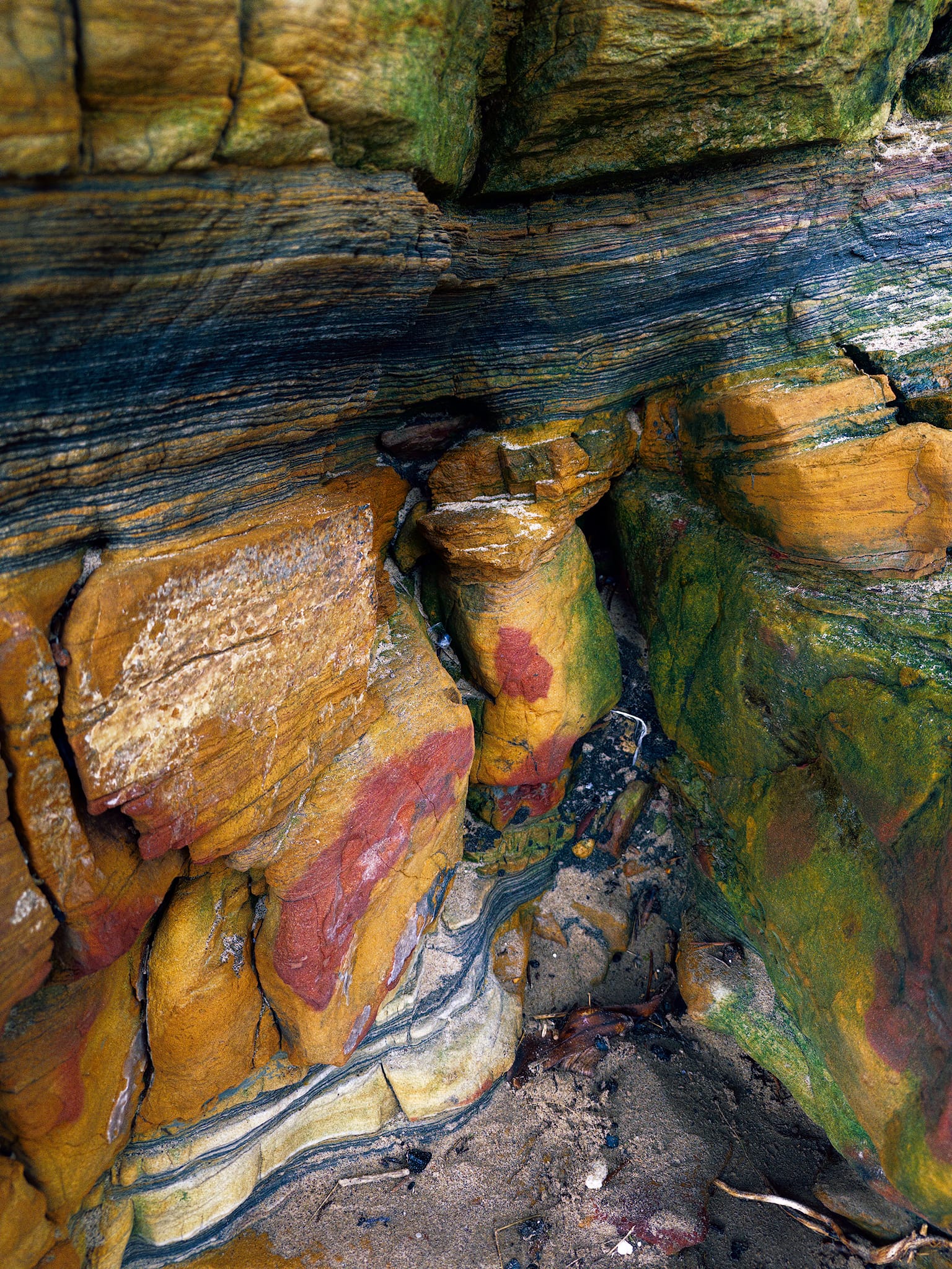 A real kaleidoscope of colour, angles, and shapes. Haematite blooms like spilled ink across ochre sandstone, beneath contorted beds liquefied by ancient seismic shock and dark carbonaceous seams. Three hundred million years of delta, swamp, and chemistry squeezed into one small nook.