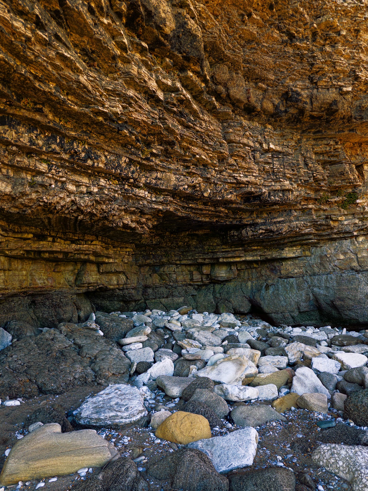 The stunning, layered cliff here is seen in cross-section. Each horizontal band shows a different layer of sediment that was deposited in an ancient, shallow sea. The thinner, finely layered lower beds are followed by thicker, more solid layers above, indicating that the conditions in the water changed over time. Near the base, there&rsquo;s a prominent overhang, which is a wave-cut notch. Over time, this overhang will likely fall off, and that’s how the boulder beach below was created. The lighter, white stones are fresher pieces of limestone that have recently fallen, while the darker, smoother ones have been shaped and polished by the waves over a longer period.
